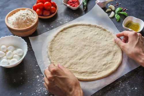 pizza dough placed on a food paper with ingredients in different bowls placed nearby