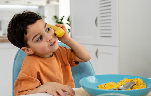A young boy holding banana as a phone while eating cereal