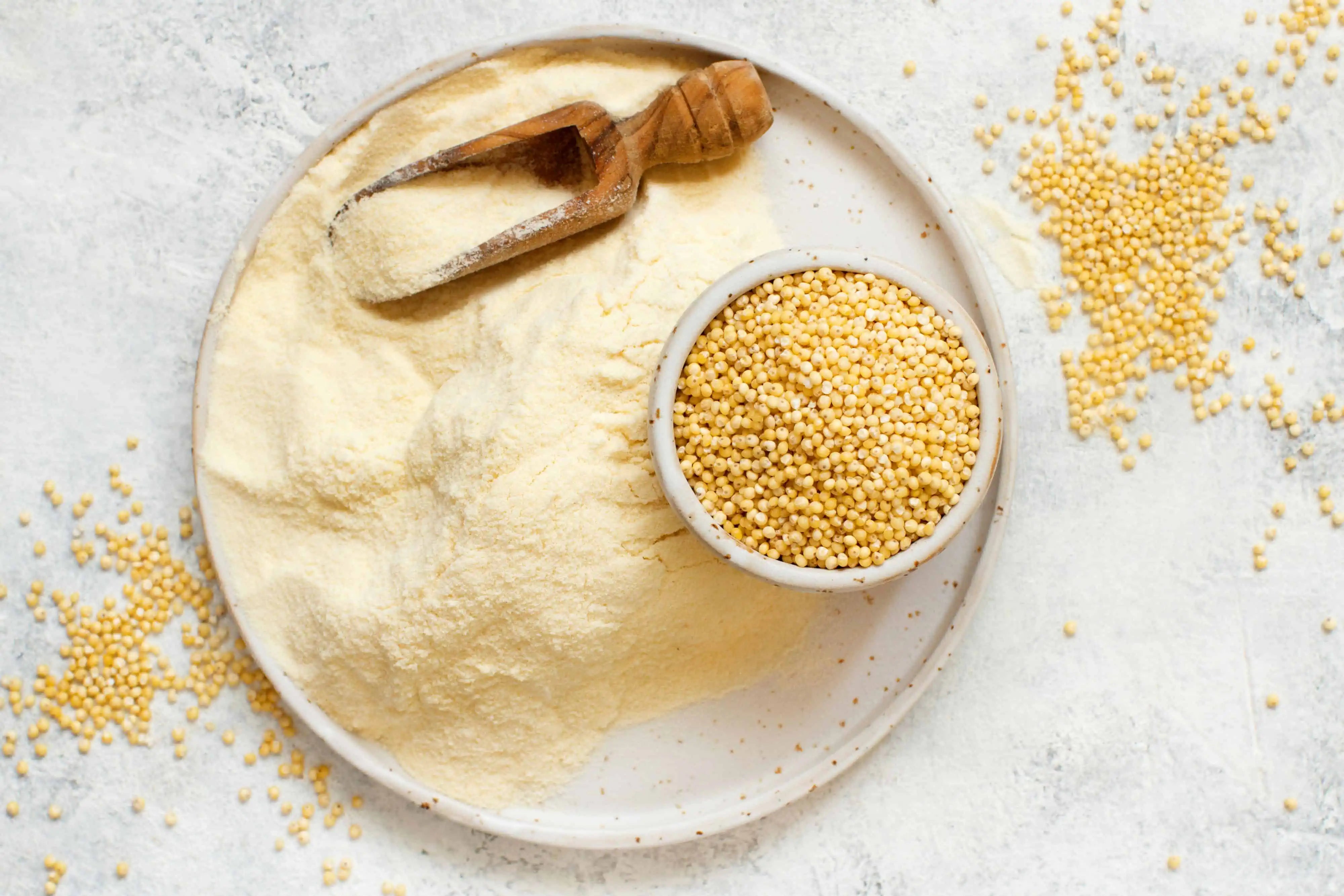 This image presents a top-down view of millet and millet flour arranged on a textured, light grey surface. A round, speckled grey plate dominates the center, partially covered with a generous mound of pale yellow millet flour. The flour isn't perfectly contained, with a soft halo extending outwards onto the surface. Resting on the flour is a small, rustic wooden scoop, also coated in the fine powder, suggesting it was recently used. Within the plate, nestled amongst the flour, is a small, white ceramic bowl brimming with whole millet grains. These grains are small, round, and a golden-yellow color, contrasting slightly with the paler flour. Scattered around the plate and on the surrounding surface are additional millet grains, creating a sense of abundance and a natural, organic feel. The lighting is soft and diffused, highlighting the textures of the flour, grains, and the plate. The overall composition is clean and minimalist, emphasizing the natural color and form of the millet in its different states – whole grain and finely ground flour. The image evokes a sense of healthy eating, baking, or a focus on natural ingredients. The background is subtly textured, adding depth without distracting from the main subject.